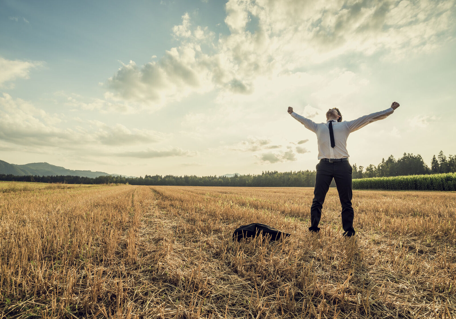 47838623 - successful businessman standing victoriously in the middle of field raising his arms in celebration and relief as he stands under majestic evening sky with his jacket lying on the floor next to him.